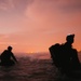 Two silhouetted figures are wading through ocean surf during twilight or nighttime conditions against a vivid orange-red sky. Both individuals are wearing tactical gear including helmets, backpacks, and what appear to be headlamps or night-vision mounts. Distant city lights are visible on the horizon, suggesting a coastal urban environment nearby.