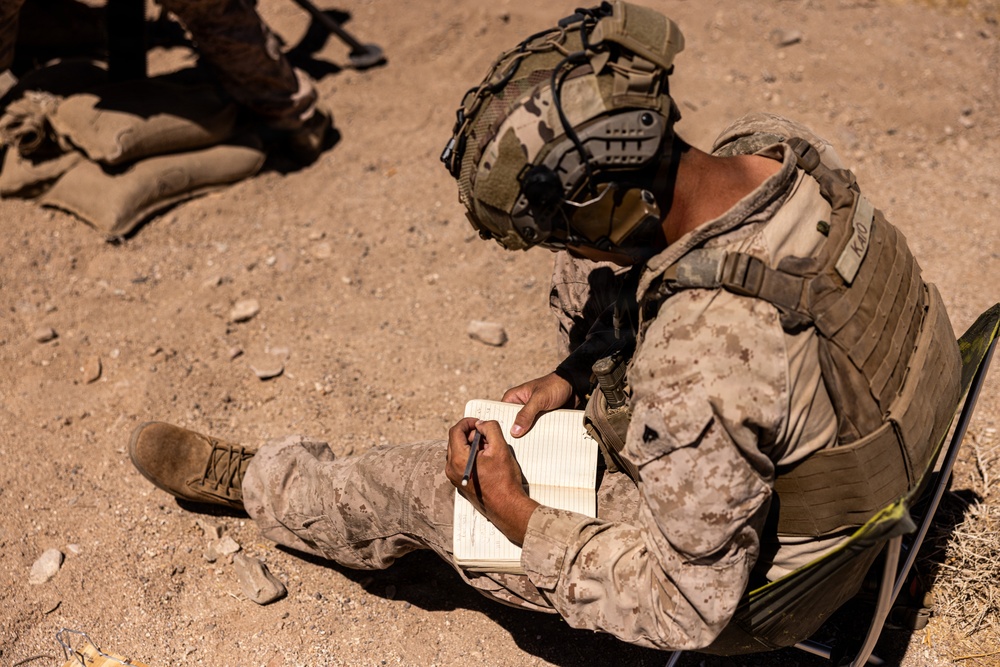 A military service member in desert camouflage (MARPAT pattern) is seated on the ground writing in a lined notebook. The soldier is fully equipped with a tactical helmet fitted with communication gear, body armor/plate carrier, and combat boots. The setting appears to be an arid, desert training or operational environment.