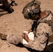 A military service member in desert camouflage (MARPAT pattern) is seated on the ground writing in a lined notebook. The soldier is fully equipped with a tactical helmet fitted with communication gear, body armor/plate carrier, and combat boots. The setting appears to be an arid, desert training or operational environment.
