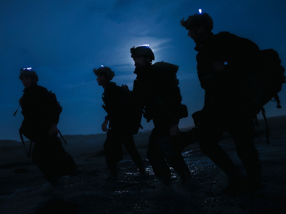 Four silhouetted figures in full tactical/military gear are moving across what appears to be a wet terrain at night. They are equipped with helmets, body armor, backpacks, and blue indicator lights visible on their helmets. A bright moon or light source is visible in the background, creating a dramatic blue-toned silhouette effect.