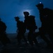 Four silhouetted figures in full tactical/military gear are moving across what appears to be a wet terrain at night. They are equipped with helmets, body armor, backpacks, and blue indicator lights visible on their helmets. A bright moon or light source is visible in the background, creating a dramatic blue-toned silhouette effect.