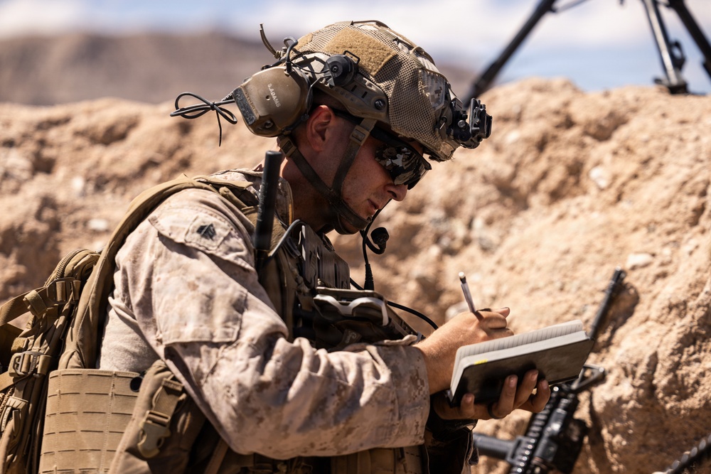 A U.S. Marine in full combat gear is writing in a small notebook in an arid, desert-like environment. The soldier is equipped with a tactical helmet, communication headset, night vision mount, body armor, and a backpack. The setting appears to be a field training or operational environment with sandy terrain and rocky berms in the background.