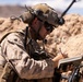 A U.S. Marine in full combat gear is writing in a small notebook in an arid, desert-like environment. The soldier is equipped with a tactical helmet, communication headset, night vision mount, body armor, and a backpack. The setting appears to be a field training or operational environment with sandy terrain and rocky berms in the background.