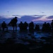 A group of approximately 10 individuals are silhouetted against a twilight sky at a beach or coastal location. Most individuals appear to be wearing helmets or headgear equipped with blue LED lights, suggesting a military, law enforcement, or specialized training context. The scene is captured during dusk or pre-dawn hours with a dramatic purple and blue sky backdrop.