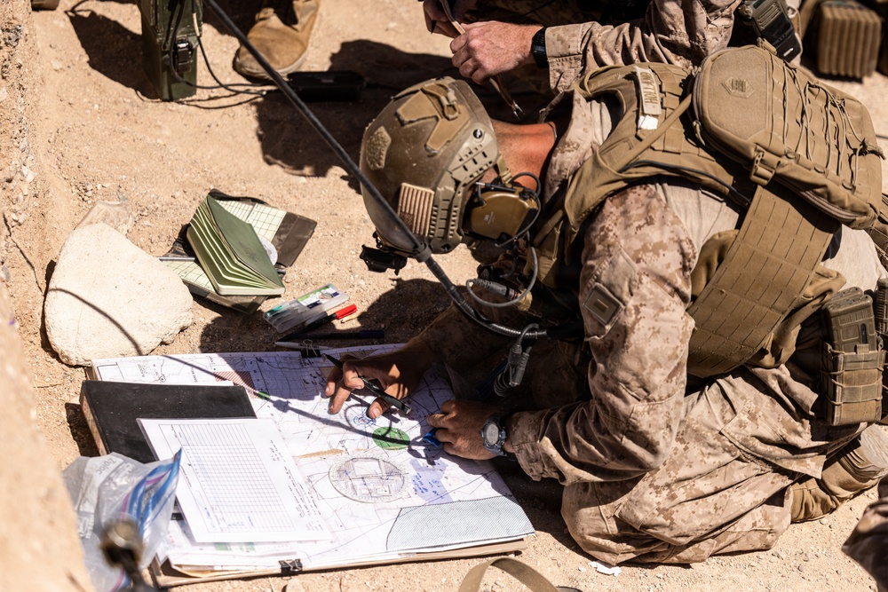 A military personnel in full combat gear is crouched on sandy ground, working with maps and navigation tools. The soldier is wearing desert camouflage (MARPAT pattern consistent with USMC), a tactical helmet with communications headset, and a plate carrier vest. They appear to be conducting map reading or fire mission planning using a compass and printed documents.