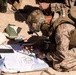 A military personnel in full combat gear is crouched on sandy ground, working with maps and navigation tools. The soldier is wearing desert camouflage (MARPAT pattern consistent with USMC), a tactical helmet with communications headset, and a plate carrier vest. They appear to be conducting map reading or fire mission planning using a compass and printed documents.
