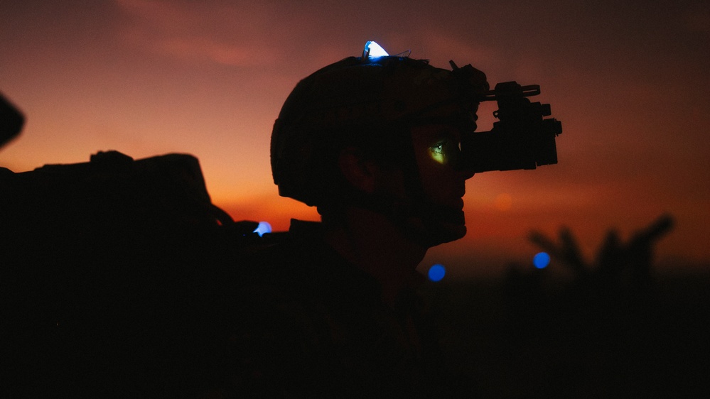 A silhouetted military personnel is photographed against a vivid orange and red twilight sky. The subject is wearing a tactical helmet equipped with night vision goggles (NVG), with a faint green glow visible through the optic lens. A small blue/white light is visible on top of the helmet, consistent with an IR or visible-spectrum identification marker.