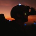 A silhouetted military personnel is photographed against a vivid orange and red twilight sky. The subject is wearing a tactical helmet equipped with night vision goggles (NVG), with a faint green glow visible through the optic lens. A small blue/white light is visible on top of the helmet, consistent with an IR or visible-spectrum identification marker.