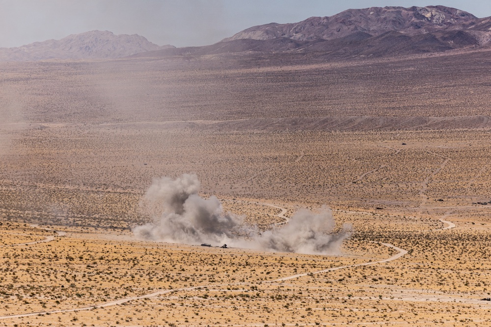 A wide desert landscape shows two large plumes of dust and smoke rising from what appears to be an explosion or detonation event near a winding dirt road. The scene is set against an arid, scrub-covered plain with mountain ranges visible in the background. The image appears to document a military exercise or controlled demolition in a remote desert environment.