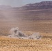 A wide desert landscape shows two large plumes of dust and smoke rising from what appears to be an explosion or detonation event near a winding dirt road. The scene is set against an arid, scrub-covered plain with mountain ranges visible in the background. The image appears to document a military exercise or controlled demolition in a remote desert environment.