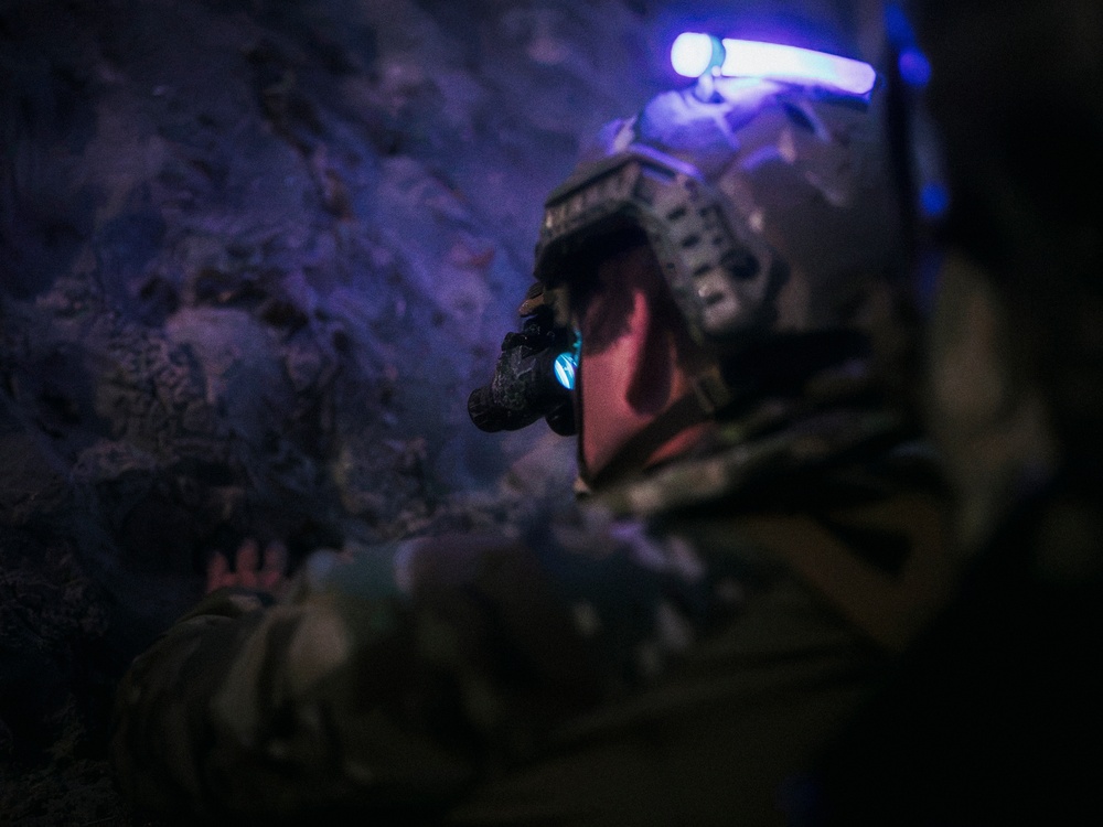 A soldier or tactical operator is photographed in low-light conditions, appearing to be inside a cave or rocky underground environment. The individual is equipped with advanced military gear including night-vision or optical devices and helmet-mounted equipment with visible blue illumination. The image is taken from a rear/side angle with shallow depth of field.