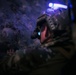 A soldier or tactical operator is photographed in low-light conditions, appearing to be inside a cave or rocky underground environment. The individual is equipped with advanced military gear including night-vision or optical devices and helmet-mounted equipment with visible blue illumination. The image is taken from a rear/side angle with shallow depth of field.