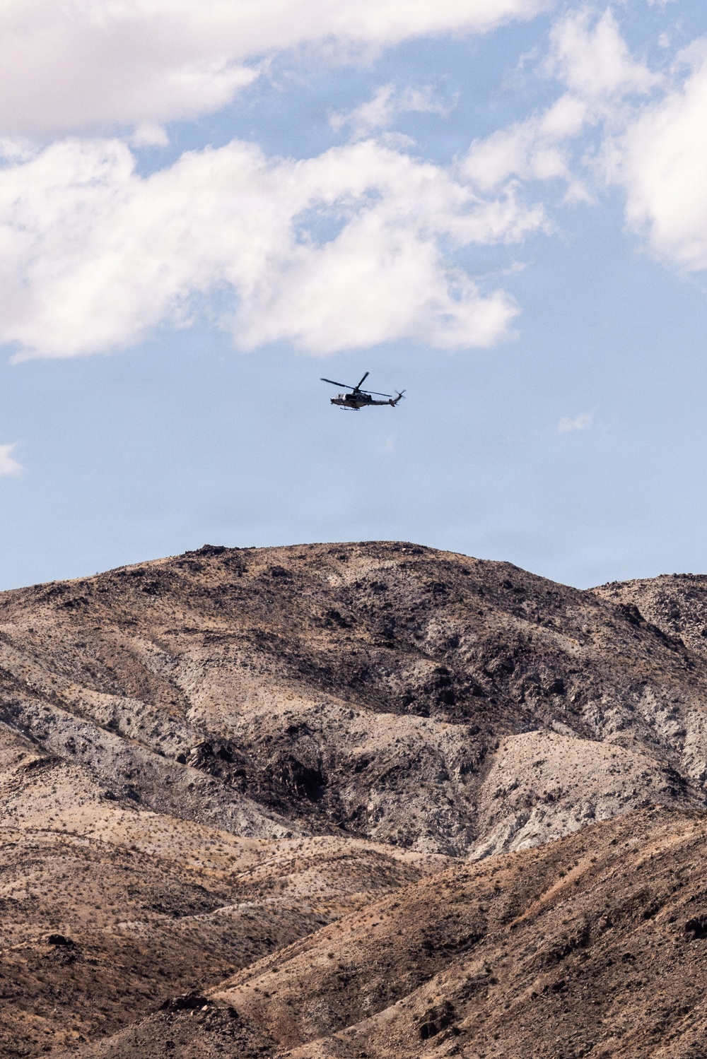 A military-style helicopter flies above a rugged, arid mountain range under a partly cloudy blue sky. The terrain below is dry and rocky, consistent with a desert environment. The helicopter appears small relative to the landscape, suggesting it is flying at a moderate altitude.