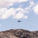 A military-style helicopter flies above a rugged, arid mountain range under a partly cloudy blue sky. The terrain below is dry and rocky, consistent with a desert environment. The helicopter appears small relative to the landscape, suggesting it is flying at a moderate altitude.