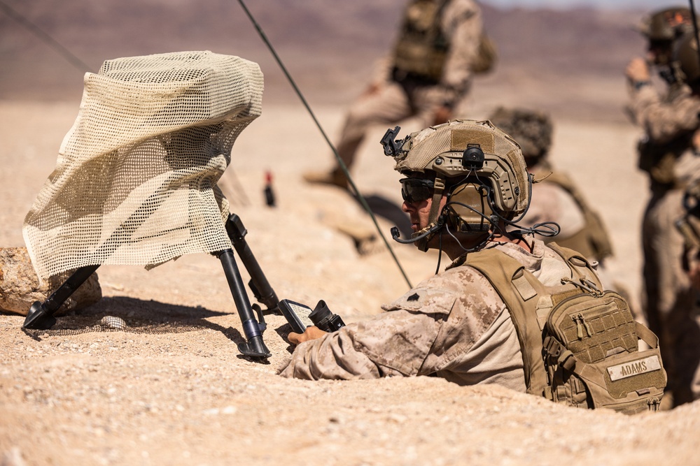 A U.S. Marine identified by the name tape 'ADAMS' is in a prone position in a desert environment, operating what appears to be a handheld tactical device. He is wearing MARPAT desert camouflage, a tactical helmet with communications equipment, and a plate carrier with a backpack. A camouflage-netted antenna or sensor on a bipod tripod is positioned nearby, with additional Marines visible in the background.