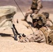 A U.S. Marine identified by the name tape 'ADAMS' is in a prone position in a desert environment, operating what appears to be a handheld tactical device. He is wearing MARPAT desert camouflage, a tactical helmet with communications equipment, and a plate carrier with a backpack. A camouflage-netted antenna or sensor on a bipod tripod is positioned nearby, with additional Marines visible in the background.