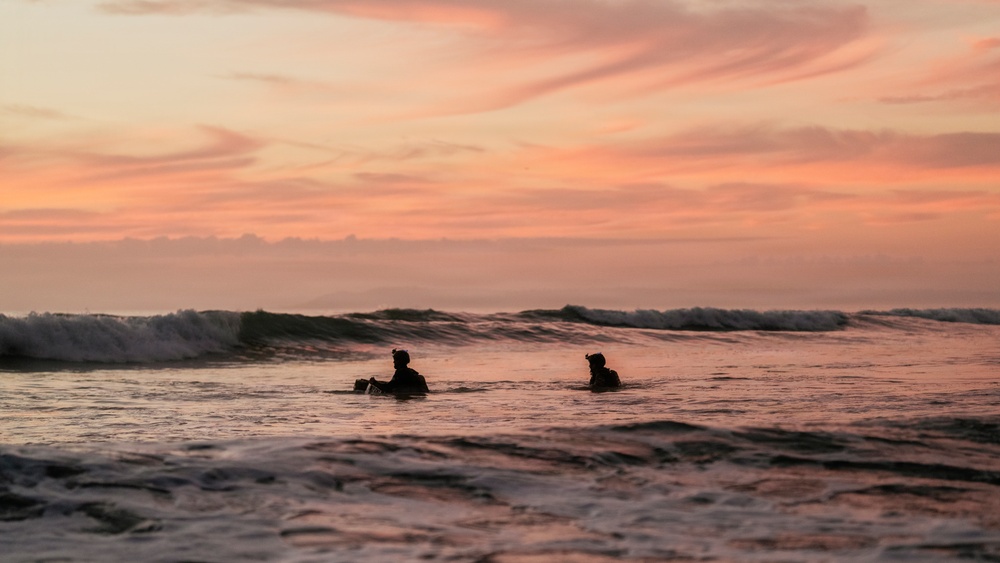 Two silhouetted figures are sitting on surfboards in the ocean during a vivid sunset. The sky is dominated by warm pink, orange, and peach tones with wispy clouds. A breaking wave is visible behind the surfers in the mid-ground.