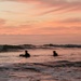 Two silhouetted figures are sitting on surfboards in the ocean during a vivid sunset. The sky is dominated by warm pink, orange, and peach tones with wispy clouds. A breaking wave is visible behind the surfers in the mid-ground.