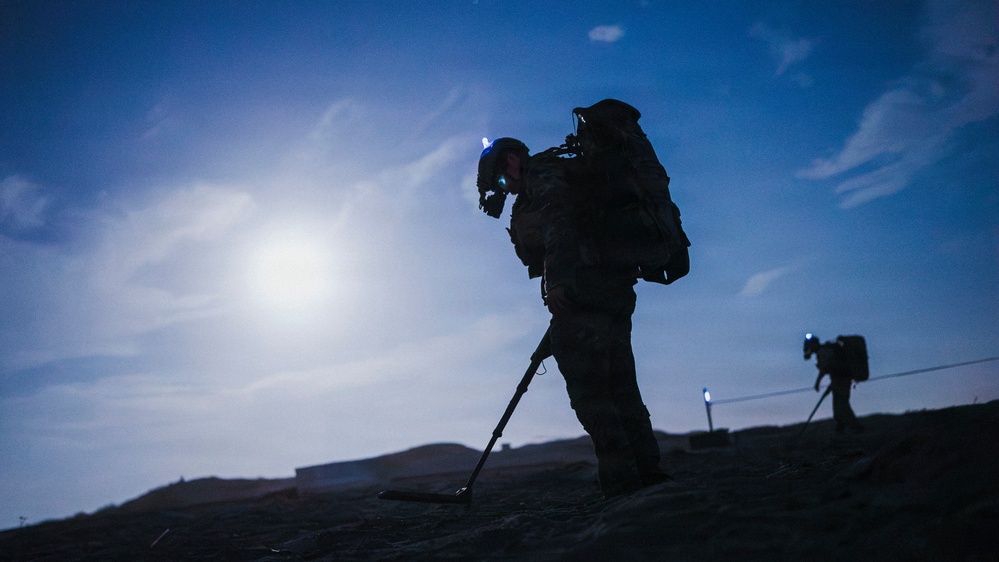 Two military personnel are silhouetted against a twilight sky while conducting what appears to be a mine-detection or route-clearance operation. The foreground figure is using a handheld metal detector or mine probe while wearing full combat kit including a large rucksack and head-mounted night-vision device. A second similarly equipped figure is visible in the background, suggesting a coordinated sweep operation.