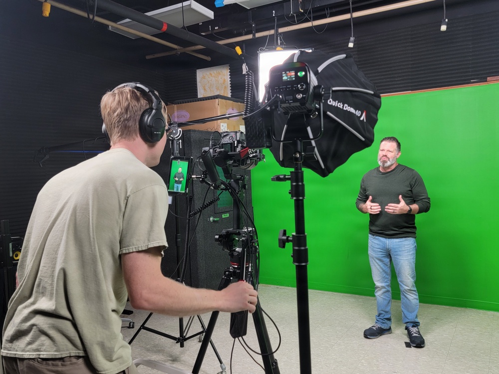 A professional video production session is taking place in a studio equipped with a green screen backdrop. A camera operator wearing headphones is filming a subject who stands in front of the green screen, gesturing as if presenting or speaking. The setup includes professional lighting equipment, a camera on a tripod, and a monitor showing the live feed.