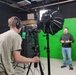 A professional video production session is taking place in a studio equipped with a green screen backdrop. A camera operator wearing headphones is filming a subject who stands in front of the green screen, gesturing as if presenting or speaking. The setup includes professional lighting equipment, a camera on a tripod, and a monitor showing the live feed.