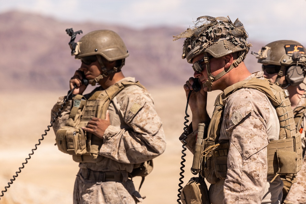 Two foreground soldiers in desert camouflage uniforms are using corded radio handsets, likely coordinating communications during a field operation or exercise. A third soldier is partially visible in the background. The arid, mountainous terrain and equipment suggest a desert training or operational environment.