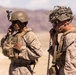 Two foreground soldiers in desert camouflage uniforms are using corded radio handsets, likely coordinating communications during a field operation or exercise. A third soldier is partially visible in the background. The arid, mountainous terrain and equipment suggest a desert training or operational environment.