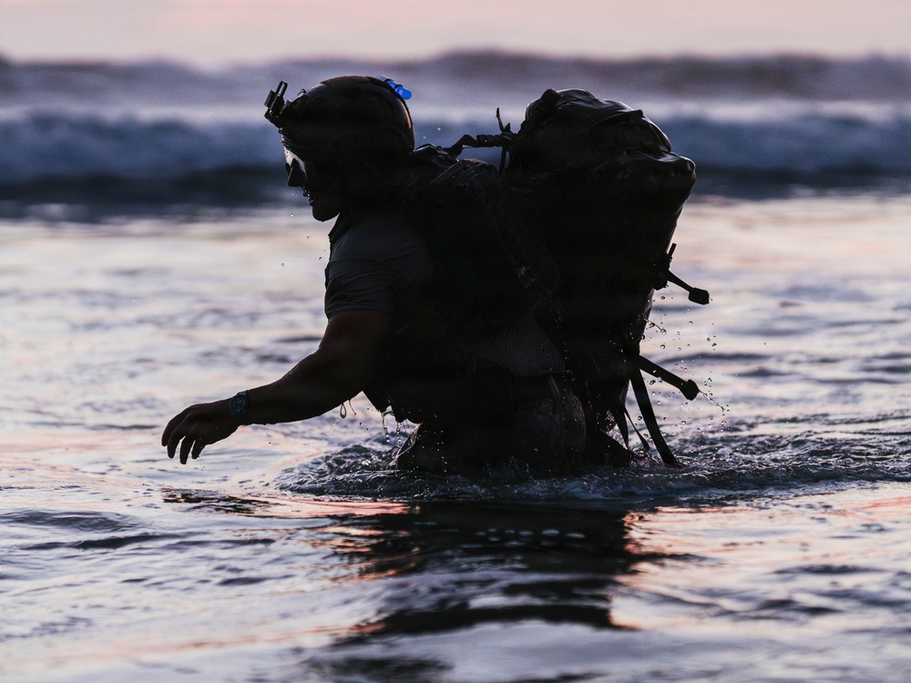 A silhouetted individual wearing tactical military gear wades through ocean surf during twilight hours. The person is equipped with a large rucksack, tactical helmet with mounted accessories, and appears to be conducting a water insertion or beach landing exercise. The dramatic low-light conditions create a stark silhouette against the pink and blue dusk sky.