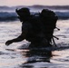 A silhouetted individual wearing tactical military gear wades through ocean surf during twilight hours. The person is equipped with a large rucksack, tactical helmet with mounted accessories, and appears to be conducting a water insertion or beach landing exercise. The dramatic low-light conditions create a stark silhouette against the pink and blue dusk sky.