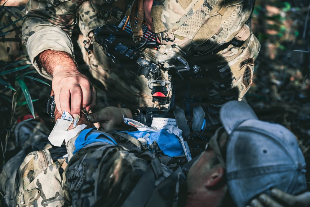 A soldier in camouflage uniform with a visible US flag patch is administering tactical combat casualty care (TCCC) to a downed individual lying on the ground. The standing soldier appears to be applying or preparing medical dressings to the casualty. The setting appears to be an outdoor wooded or vegetated environment, consistent with a field training exercise or operational scenario.