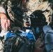 A soldier in camouflage uniform with a visible US flag patch is administering tactical combat casualty care (TCCC) to a downed individual lying on the ground. The standing soldier appears to be applying or preparing medical dressings to the casualty. The setting appears to be an outdoor wooded or vegetated environment, consistent with a field training exercise or operational scenario.