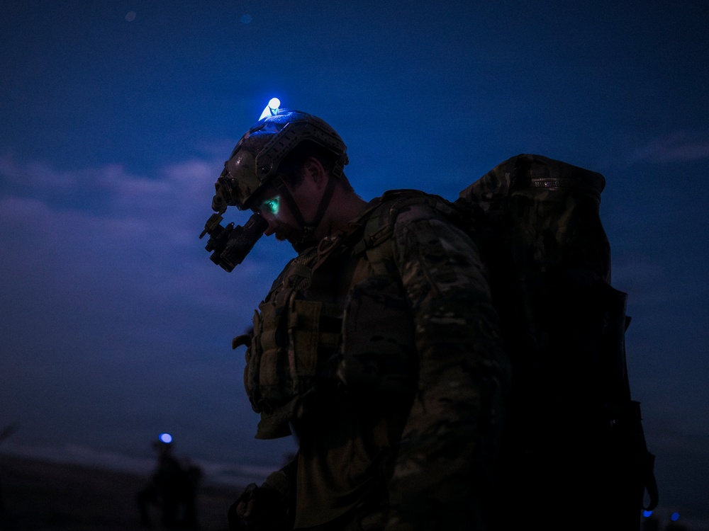 A heavily equipped soldier is photographed at night wearing full combat gear including a helmet-mounted night vision device and carrying a large rucksack. A blue indicator light (likely an IR/visible strobe or helmet light) is illuminated on top of the helmet, and green light emanates from the night vision optics. At least one additional soldier is visible in the background, also with a blue light.