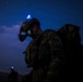 A heavily equipped soldier is photographed at night wearing full combat gear including a helmet-mounted night vision device and carrying a large rucksack. A blue indicator light (likely an IR/visible strobe or helmet light) is illuminated on top of the helmet, and green light emanates from the night vision optics. At least one additional soldier is visible in the background, also with a blue light.