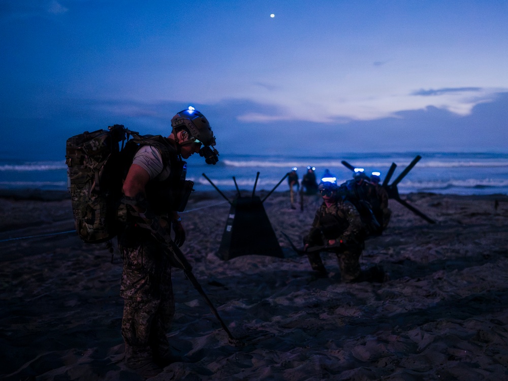 Military personnel in full tactical gear are conducting a nighttime beach operation, likely a training exercise. Multiple soldiers wearing helmets with blue IR/LED lights are carrying heavy rucksacks and navigating beach obstacles in low-light conditions. The scene is set at dusk or dawn with ocean waves visible in the background.