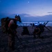 Military personnel in full tactical gear are conducting a nighttime beach operation, likely a training exercise. Multiple soldiers wearing helmets with blue IR/LED lights are carrying heavy rucksacks and navigating beach obstacles in low-light conditions. The scene is set at dusk or dawn with ocean waves visible in the background.