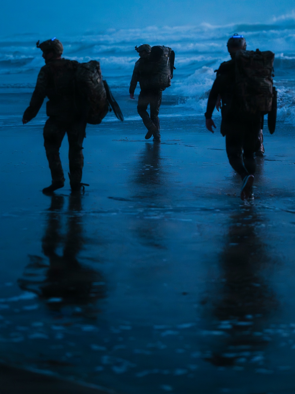 Three heavily equipped military personnel are walking toward ocean waves on a wet beach during twilight or pre-dawn hours. The figures are silhouetted against a blue-toned sky and surf, carrying large tactical rucksacks and wearing combat gear. Their reflections are visible on the wet sand, suggesting a maritime insertion or beach training exercise.