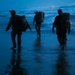 Three heavily equipped military personnel are walking toward ocean waves on a wet beach during twilight or pre-dawn hours. The figures are silhouetted against a blue-toned sky and surf, carrying large tactical rucksacks and wearing combat gear. Their reflections are visible on the wet sand, suggesting a maritime insertion or beach training exercise.