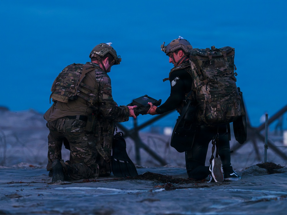 Two individuals in military/tactical gear are kneeling on a wet beach surface during twilight or pre-dawn hours. They appear to be exchanging or examining a flat device, possibly a tablet or equipment case. One person is wearing full camouflage uniform while the other is in a wetsuit with tactical vest and large rucksack.