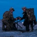 Two individuals in military/tactical gear are kneeling on a wet beach surface during twilight or pre-dawn hours. They appear to be exchanging or examining a flat device, possibly a tablet or equipment case. One person is wearing full camouflage uniform while the other is in a wetsuit with tactical vest and large rucksack.