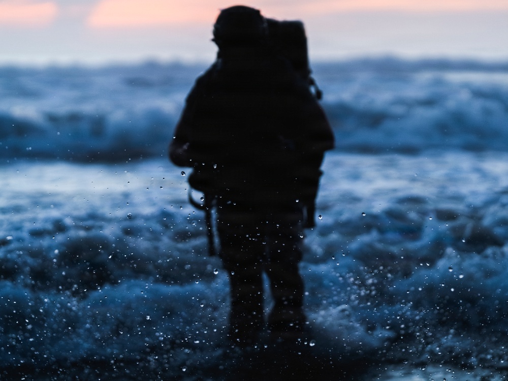 A heavily silhouetted figure, appearing to be a soldier or tactical operator with full gear and backpack, stands in shallow ocean surf during twilight or dawn. The image is shot through a wet lens, creating water droplets in the foreground that add a dramatic, immersive quality. The dark blue tones of the churning waves contrast with a faint pink-orange sky on the horizon.