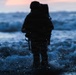 A heavily silhouetted figure, appearing to be a soldier or tactical operator with full gear and backpack, stands in shallow ocean surf during twilight or dawn. The image is shot through a wet lens, creating water droplets in the foreground that add a dramatic, immersive quality. The dark blue tones of the churning waves contrast with a faint pink-orange sky on the horizon.