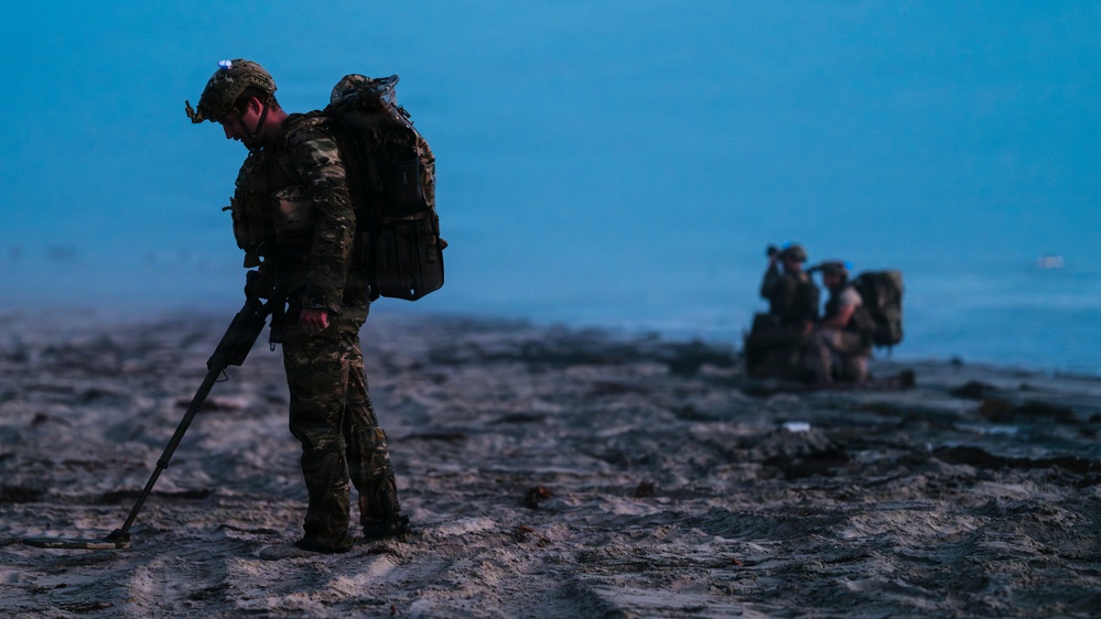 Military personnel are conducting operations on a beach at dusk or dawn in low-light conditions. The foreground soldier is using what appears to be a mine detector or probing rod while scanning the sandy terrain. Additional personnel are visible in the background, one appearing to use optics or binoculars.