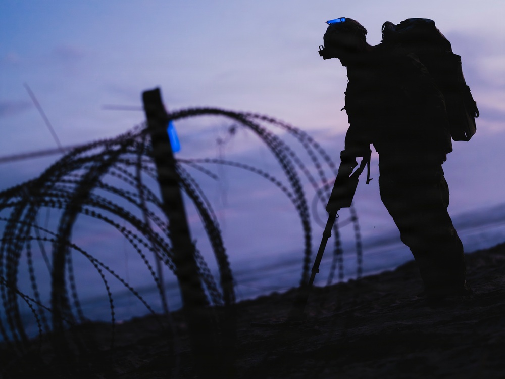 A silhouetted soldier in full combat gear is photographed at dusk or dawn near coils of concertina/razor wire. The figure appears to be conducting a patrol or mine-detection sweep, holding what appears to be a long-handled detection instrument. The dramatic twilight sky creates a high-contrast silhouette effect, rendering most details as dark outlines.