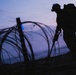 A silhouetted soldier in full combat gear is photographed at dusk or dawn near coils of concertina/razor wire. The figure appears to be conducting a patrol or mine-detection sweep, holding what appears to be a long-handled detection instrument. The dramatic twilight sky creates a high-contrast silhouette effect, rendering most details as dark outlines.