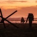 A dramatic silhouette image showing military personnel on a beach at sunset or sunrise, with World War II-style beach obstacles (Czech hedgehogs) and barbed wire in the foreground. Two figures in full combat gear with rifles and backpacks are visible advancing toward the waterline. The scene strongly evokes D-Day or amphibious assault imagery, either historical recreation or military exercise.