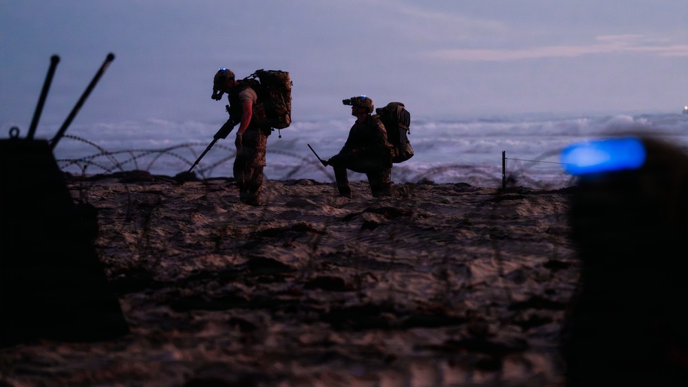 Two silhouetted military personnel are operating on a beach at dusk or dawn, equipped with tactical gear and backpacks. One figure appears to be using a metal detector or probe while the other uses optical/night-vision equipment. Concertina wire and what appears to be a defensive position are visible in the foreground, suggesting a beach security or EOD (explosive ordnance disposal) operation.