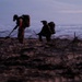 Two silhouetted military personnel are operating on a beach at dusk or dawn, equipped with tactical gear and backpacks. One figure appears to be using a metal detector or probe while the other uses optical/night-vision equipment. Concertina wire and what appears to be a defensive position are visible in the foreground, suggesting a beach security or EOD (explosive ordnance disposal) operation.