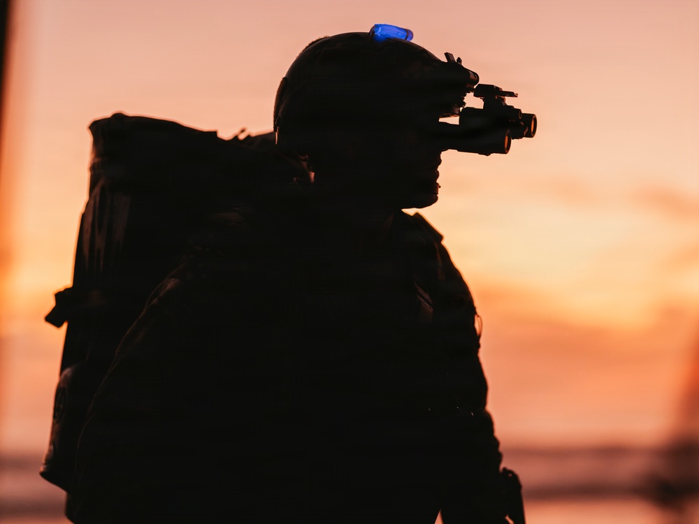 A silhouetted figure wearing military-style tactical gear is photographed against a vivid orange and pink sunset sky. The subject is equipped with night vision goggles mounted on a helmet and carries a large backpack. The image is a dramatic backlit silhouette composition with a single blue light visible on the helmet.