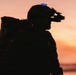 A silhouetted figure wearing military-style tactical gear is photographed against a vivid orange and pink sunset sky. The subject is equipped with night vision goggles mounted on a helmet and carries a large backpack. The image is a dramatic backlit silhouette composition with a single blue light visible on the helmet.
