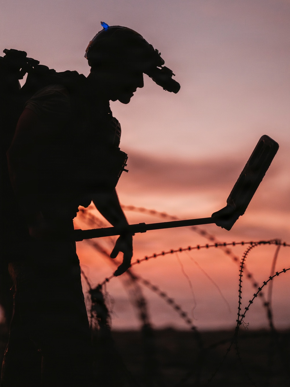 A silhouetted military or security personnel figure is photographed against a vivid orange-pink sunset sky. The individual is equipped with a tactical helmet fitted with night-vision goggles and a blue indicator light, and is holding what appears to be a metal detector or mine-detection device. Coils of razor/concertina wire are visible in the foreground, suggesting a secured perimeter or demining operation.